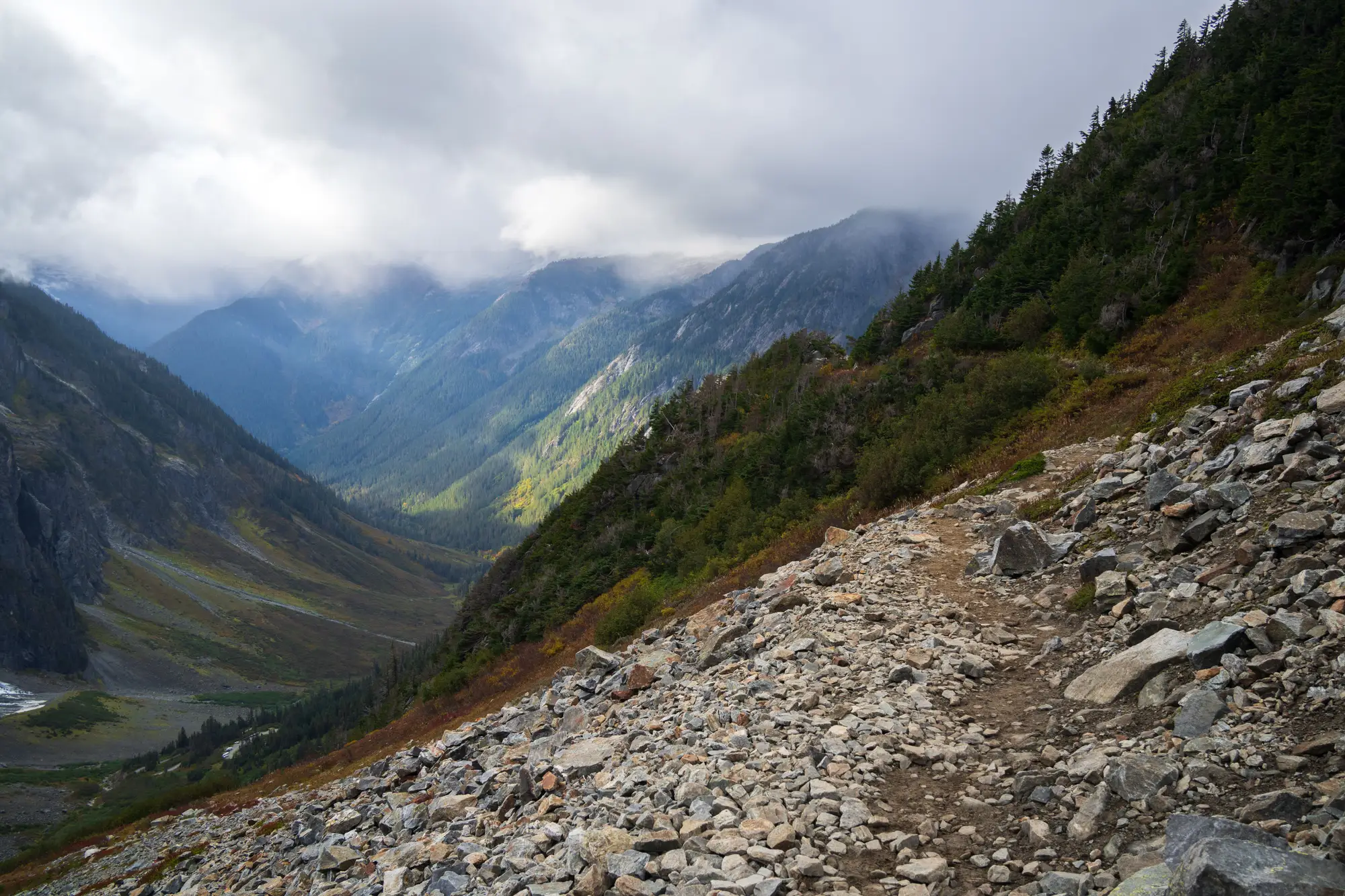 A rocky trail crossing a scree slope above a forested valley, low cloud on the ridges